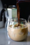 A glass filled with iced milk is being poured over with coffee, creating a swirl of mixed colors. The background features a blurred silver espresso maker with a black handle. The surface is a light-colored countertop.