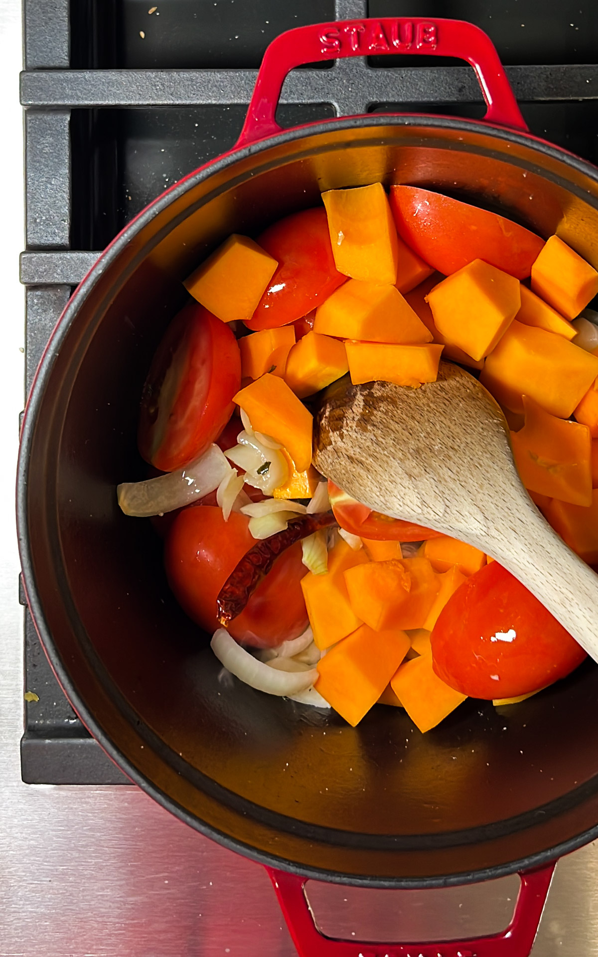Una olla roja con verduras picadas para sopa de calabaza y una cuchara de madera sobre una estufa.
