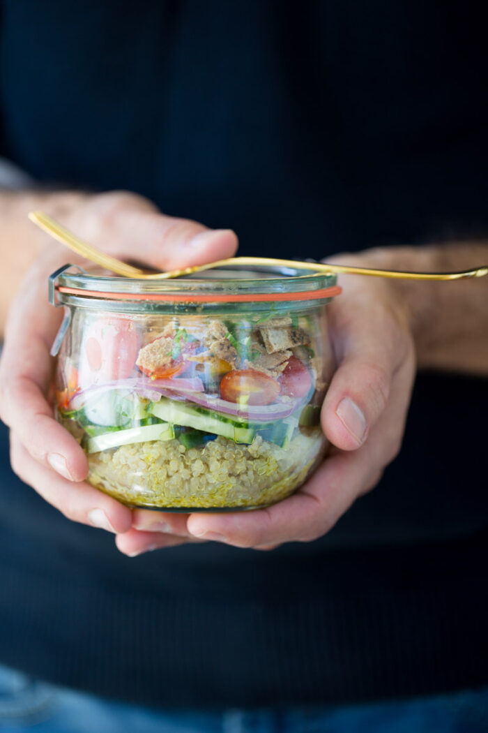 Una persona sostiene un frasco de vidrio lleno de una vibrante ensalada mediterránea de quinoa. Incluye capas de quinoa, pepinos en rodajas, tomates cherry y pollo a la parrilla. Un tenedor dorado está escondido debajo de la tapa. El fondo está difuminado, lo que enfatiza el colorido contenido de esta delicia saludable.