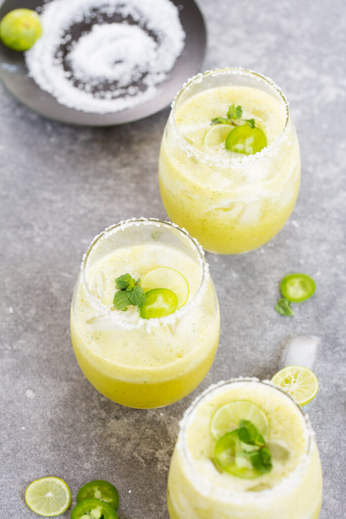 Tres vasos de margaritas frías de color amarillo verdoso, adornados con rodajas de lima, rodajas de jalapeño y menta fresca. Los bordes están cubiertos de sal. Un plato negro al fondo contiene una lima y sal. Rodajas circulares de lima y jalapeño se encuentran esparcidas sobre la superficie gris, enmarcando a la perfección la escena de la margarita.