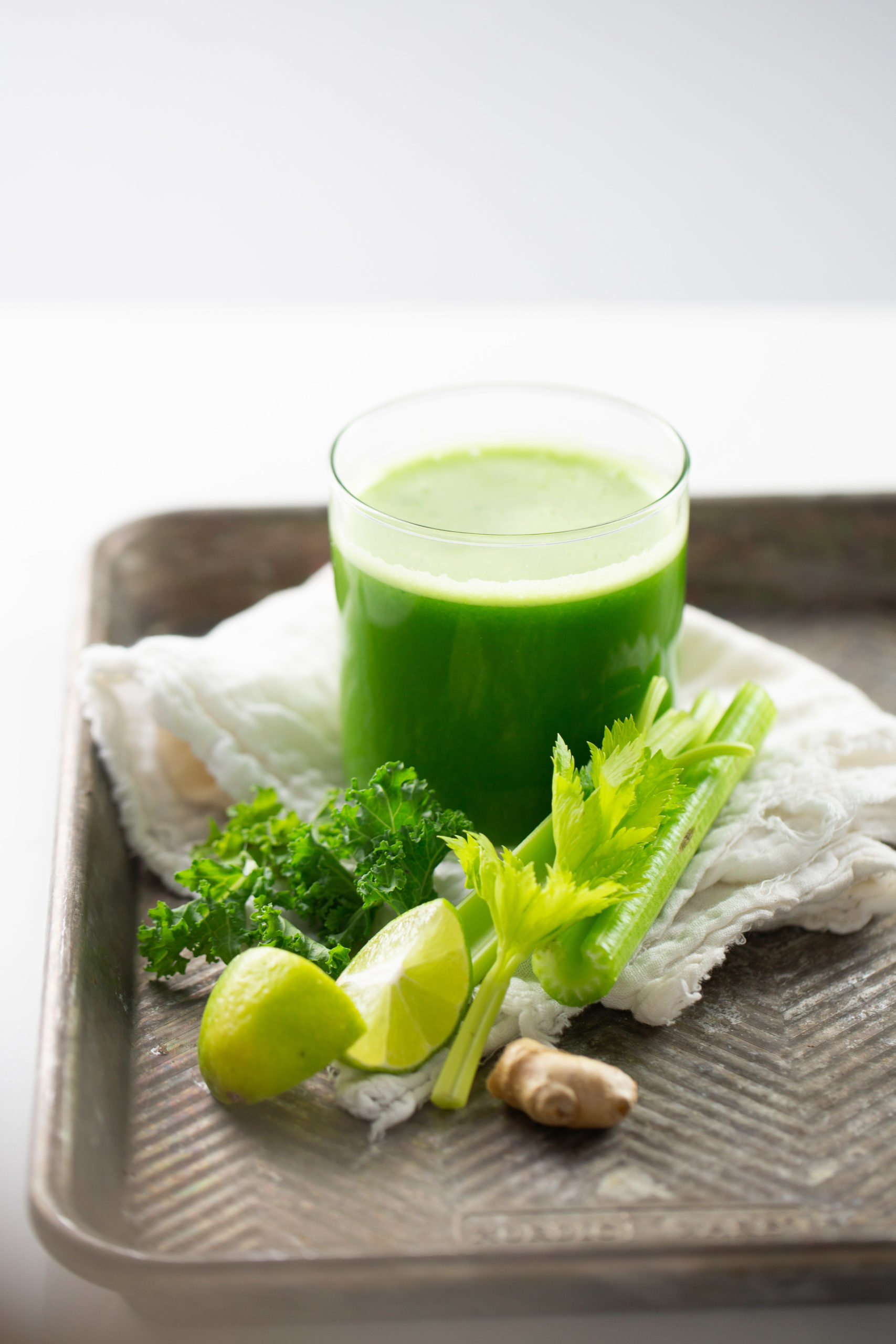 jugo verde hecho en casa en una charola con limón, apio, kale y jengibre a un lado.