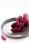 A silver tray holds several fresh beets with long, vibrant pink stems and leaves. One beet is sliced, revealing its deep red interior. The tray is placed on a white surface, and a small beet slice rests nearby, adding a touch of color contrast.