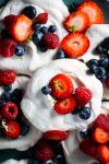 Close-up of vegan pavlova nests topped with fresh fruit. Each meringue is adorned with vibrant strawberries, blueberries, and raspberries. The meringues have a light, airy texture, contrasting with the colorful fruits. The composition highlights the freshness and natural colors.