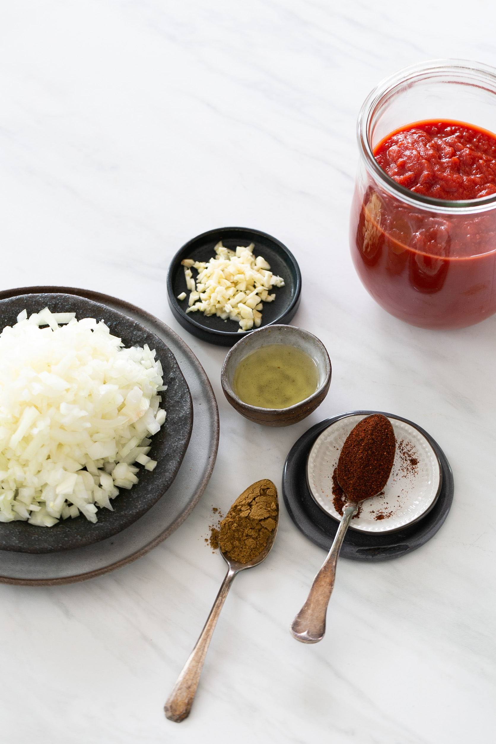 Sobre una encimera de mármol, un plato con cebollas finamente picadas, un cuenco pequeño con ajo picado y un cuenco pequeño con jengibre picado se encuentran junto a un recipiente con salsa de tomate. Una cucharada de chile en polvo y comino molido insinúan los sabores perfectos para una slasa para enchiladas con chipotle. Dos cuencos negros y uno gris contienen los ingredientes.