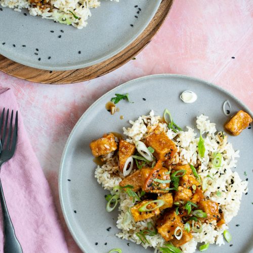 Un plato de arroz y tofu sobre fondo rosa, cubierto con tofu crujiente de naranja.