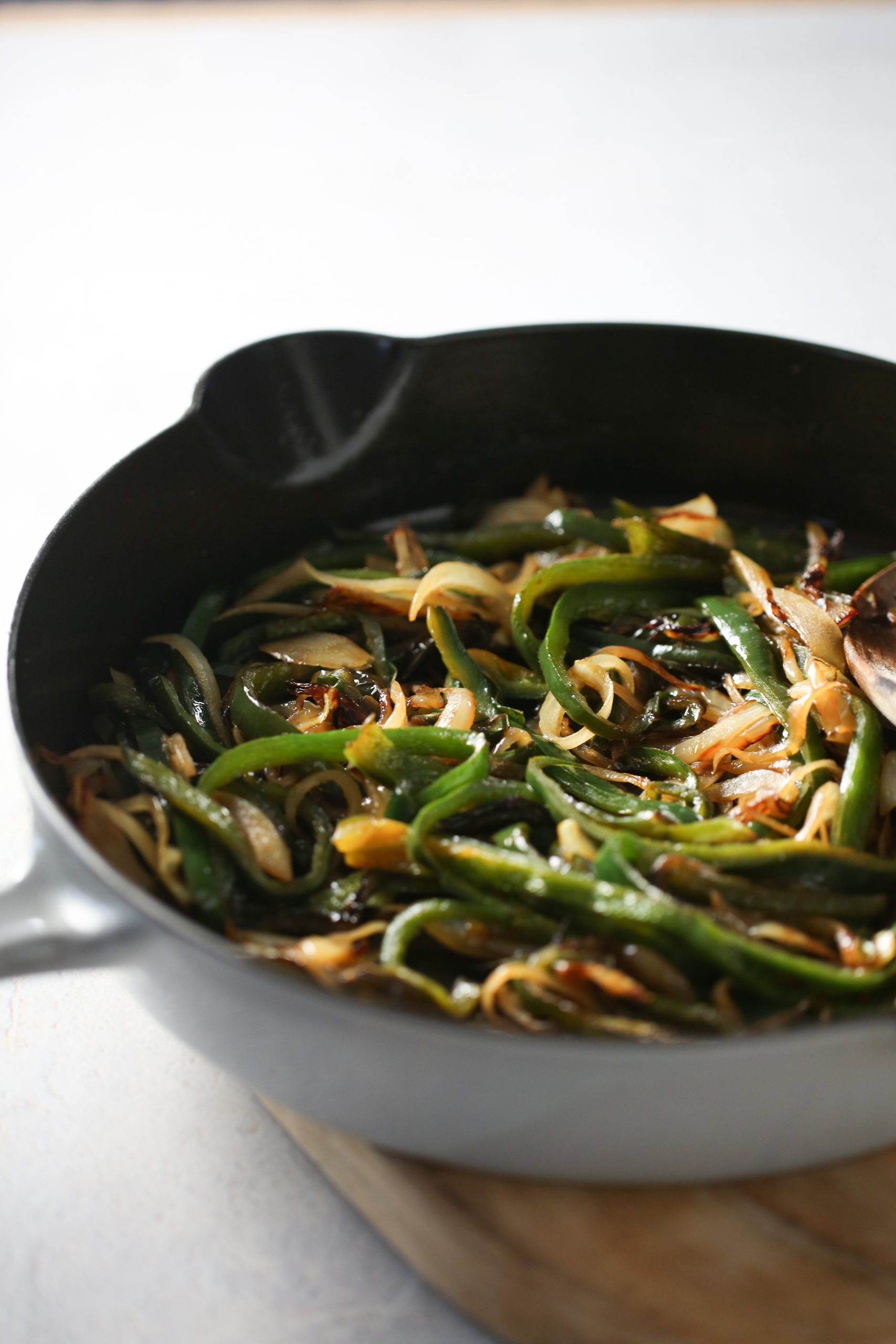 Primer plano de una sartén negra sobre una tabla de madera que contienepoblanos y cebollas salteados, que recuerdan a las rajas con crema veganas. Las verduras están ligeramente doradas y caramelizadas, con una cuchara de madera parcialmente visible en el lado derecho de la sartén. El fondo está suavemente difuminado, lo que resalta el contenido de la sartén.