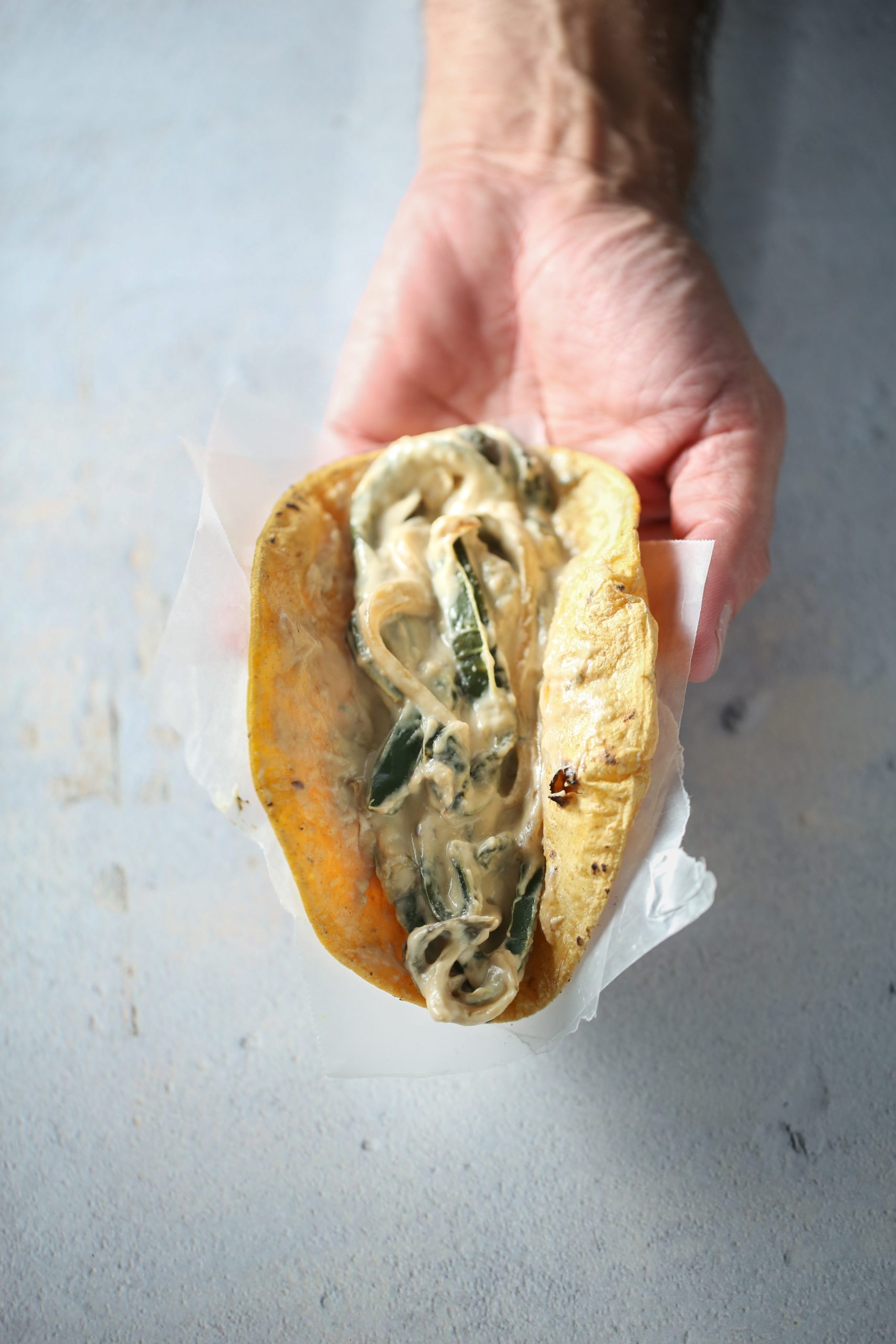 Primer plano de una mano que sostiene un taco abierto envuelto en papel encerado. El taco, relleno de rajas con crema vegana, tiene una tortilla de maíz amarilla que contiene cebollas y pimientos salteados cremosos. El fondo revela una superficie clara y texturizada.