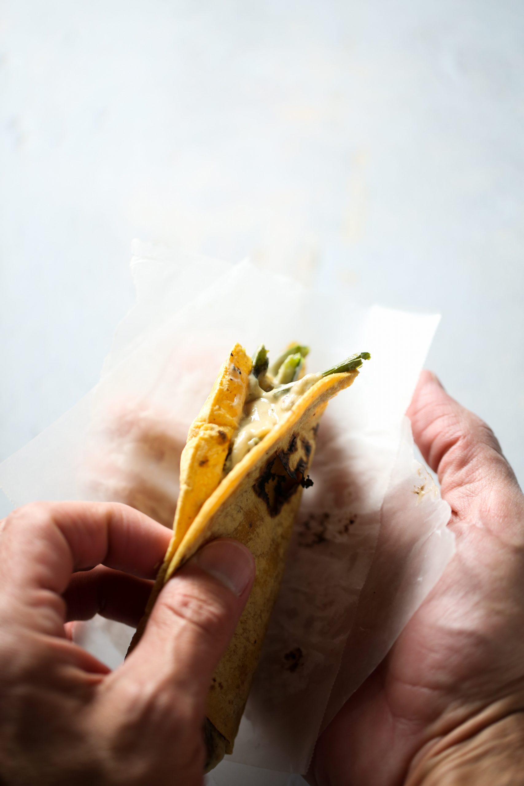 Un par de manos que sostienen un taco parcialmente envuelto. El taco, relleno de rajas con crema vegana, vegetales verdes y queso derretido, parece estar hecho con una tortilla de maíz. El papel de envolver es semitranslúcido y ligeramente grasoso debido al relleno del taco. El fondo es una superficie gris clara y lisa.