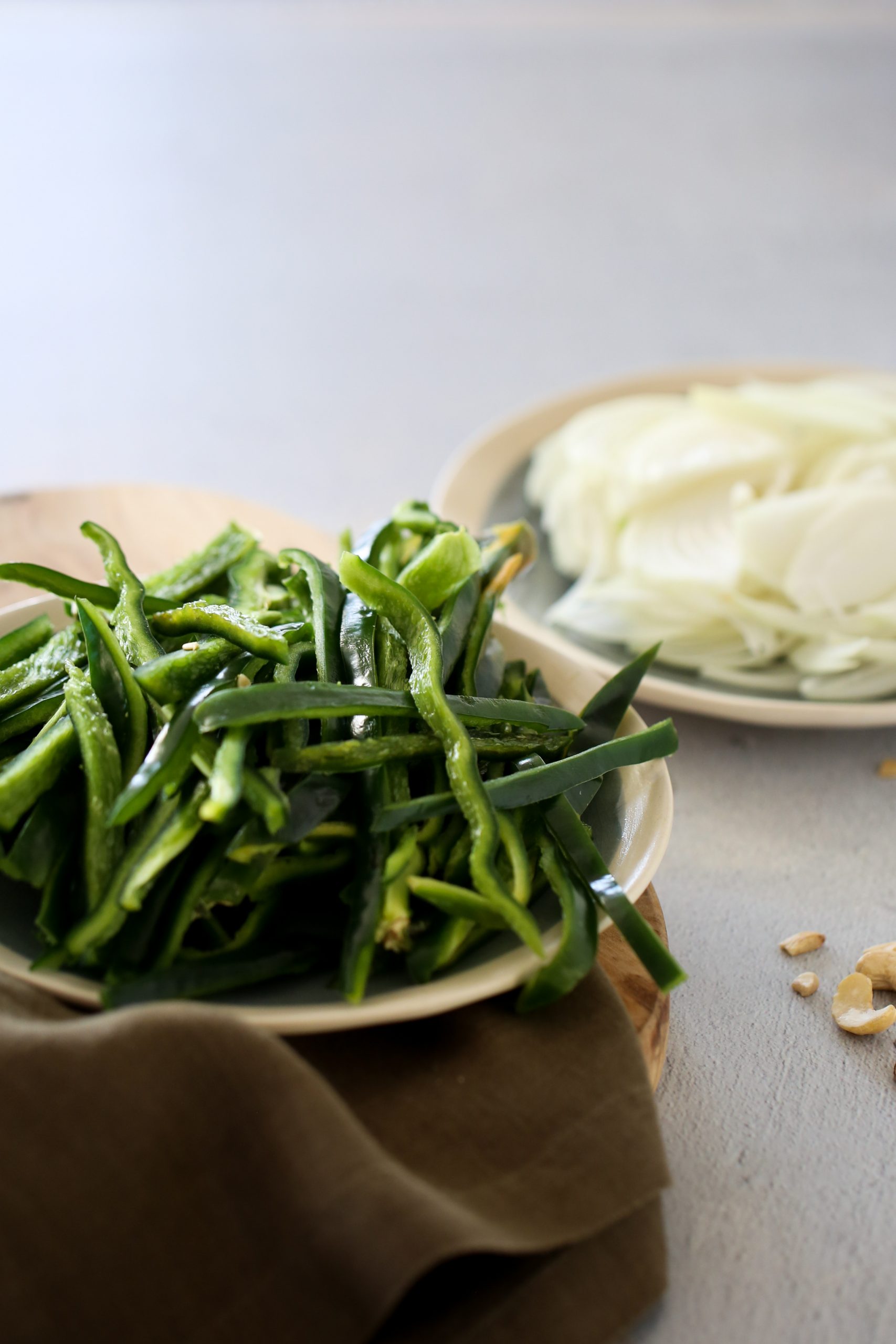 En primer plano, sobre una superficie de madera, se encuentra un plato con poblanosverdes cortados en rodajas y junto a él una servilleta de tela marrón. Al fondo, hay otro plato con cebollas cortadas en rodajas, perfectas para preparar unas deliciosas rajas con crema veganas. El entorno parece ser una cocina o un comedor.