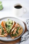 Three tacos filled with Mexican green beans and various crumbled toppings sit on a gray plate. A white mug filled with black coffee is in the background, and a patterned cloth drapes partially over the wooden surface beside the plate. The setting is bright with natural lighting.