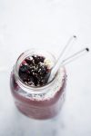 A top view of a mason jar filled with a vibrant acai smoothie, topped with blueberries, shredded coconut, and sprinkled sesame seeds. Two clear glass straws are inserted into the smoothie. The jar sits on a light, textured surface.