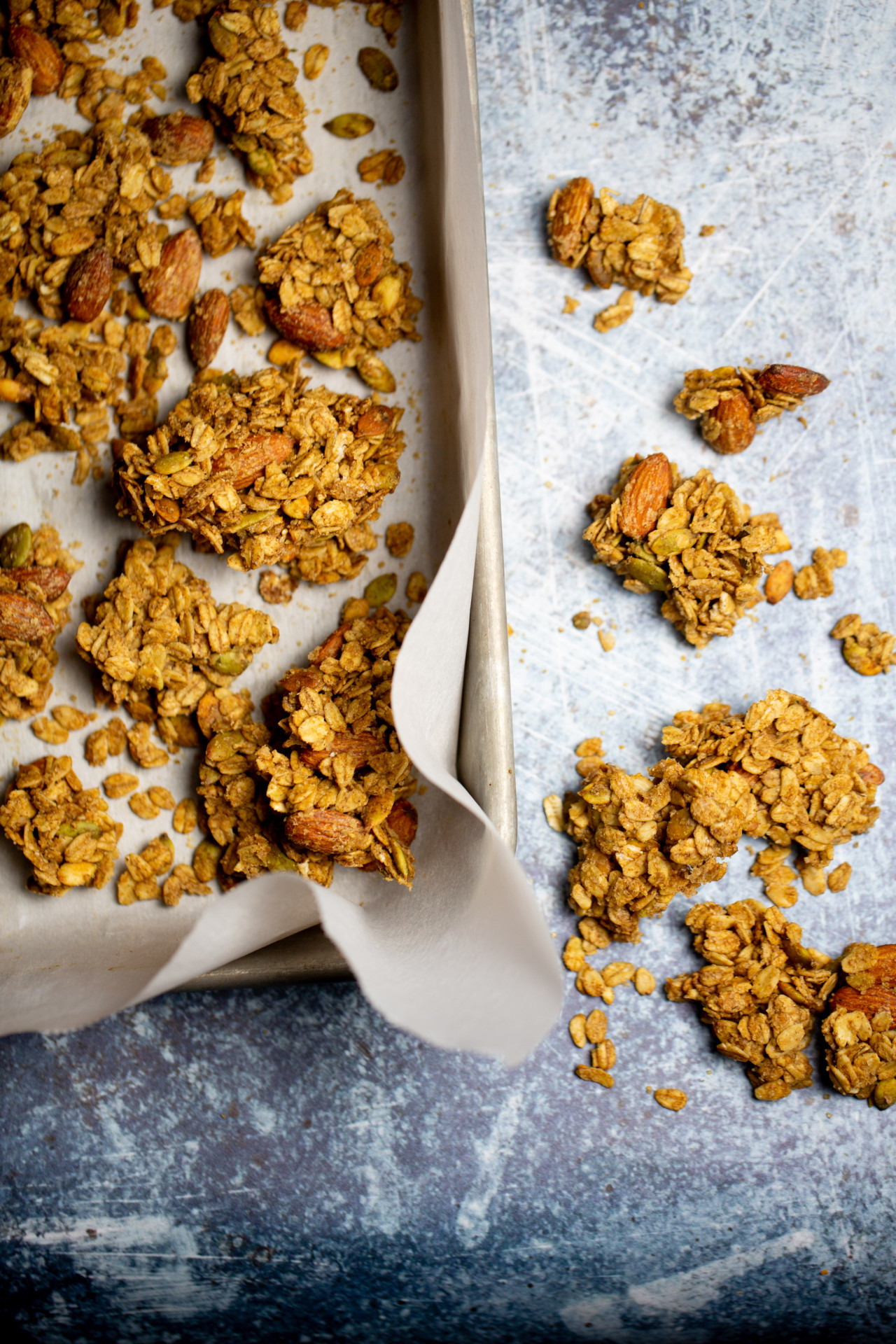 Primer plano de una bandeja para hornear forrada con papel de pergamino, llena de racimos de trozos de granola de color marrón dorado. La granola incluye almendras y semillas de girasol visibles. Algunos racimos se derraman sobre la superficie gris texturizada junto a la bandeja, lo que crea una presentación rústica.