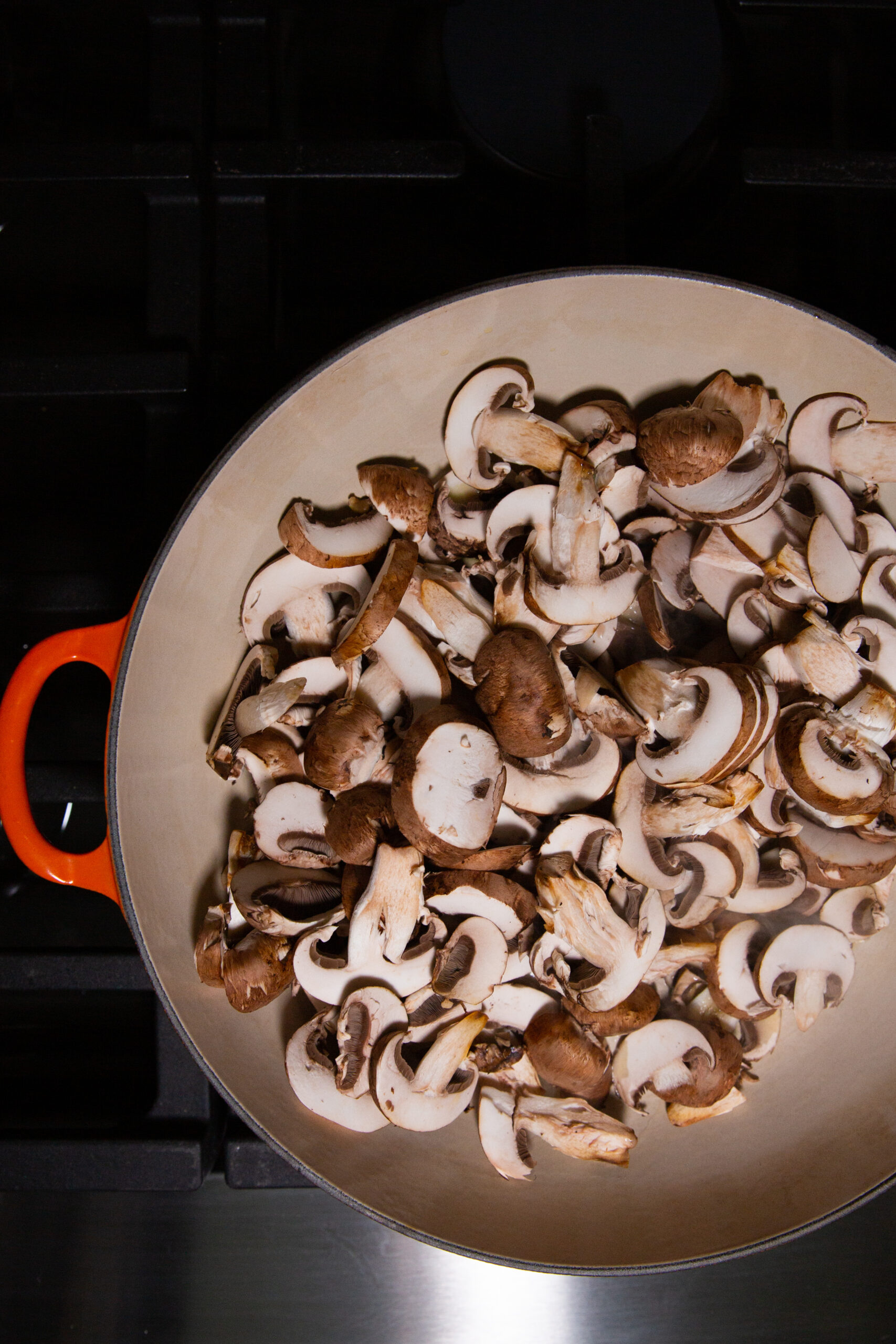 Una olla grande con un asa naranja está llena de champiñones marrones cortados en rodajas. La olla se coloca sobre la hornilla de una estufa, lista para cocinar. Los hongos varían de tamaño y están distribuidos uniformemente en el interior de color claro de la olla, ideal para un delicioso plato de pasta.