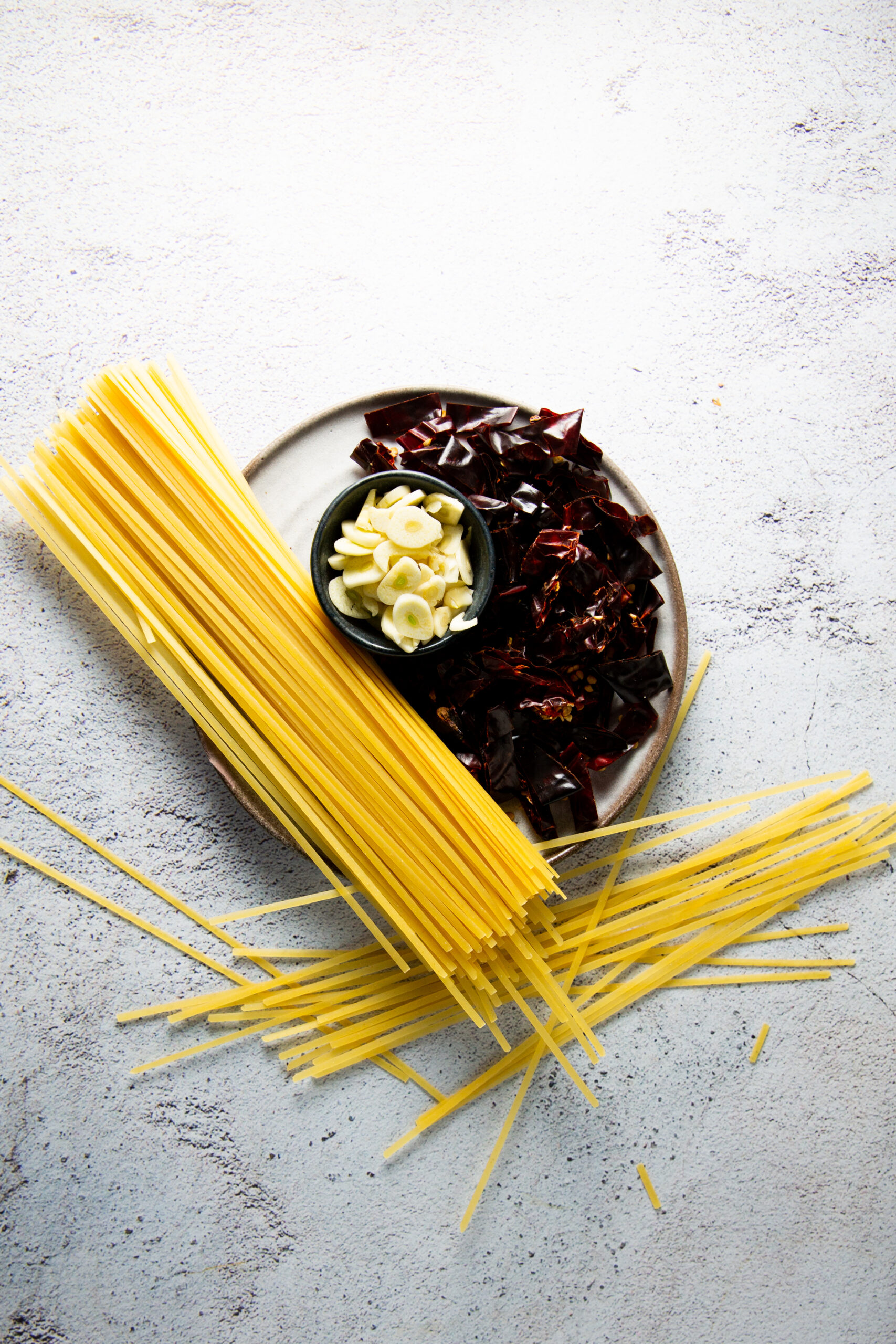 Un plato contiene ingredientes para un plato de pasta: espaguetis secos, dientes de ajo en rodajas finas en un tazón negro pequeño y trozos de chile guajillo seco. Los ingredientes están esparcidos sobre una superficie clara y texturizada.