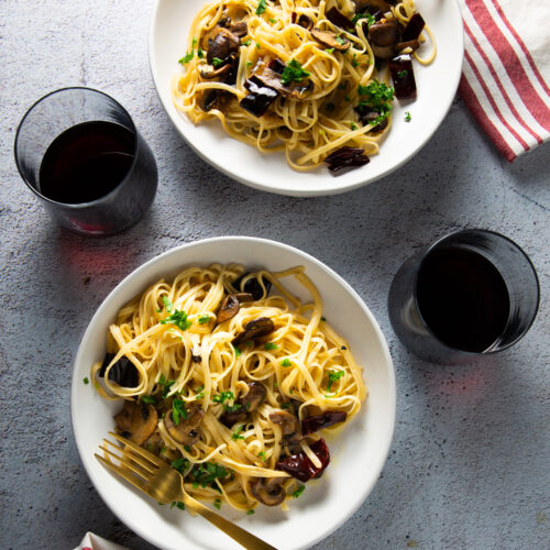 Sobre una superficie de color claro se encuentran dos cuencos blancos de pasta con champiñones y hierbas. Cada cuenco tiene un tenedor, uno dorado y otro plateado. Junto a los cuencos hay dos copas llenas de vino tinto. Junto a cada cuenco se coloca una servilleta de rayas blancas y rojas, que añade un toque de elegancia a la escena bien iluminada.