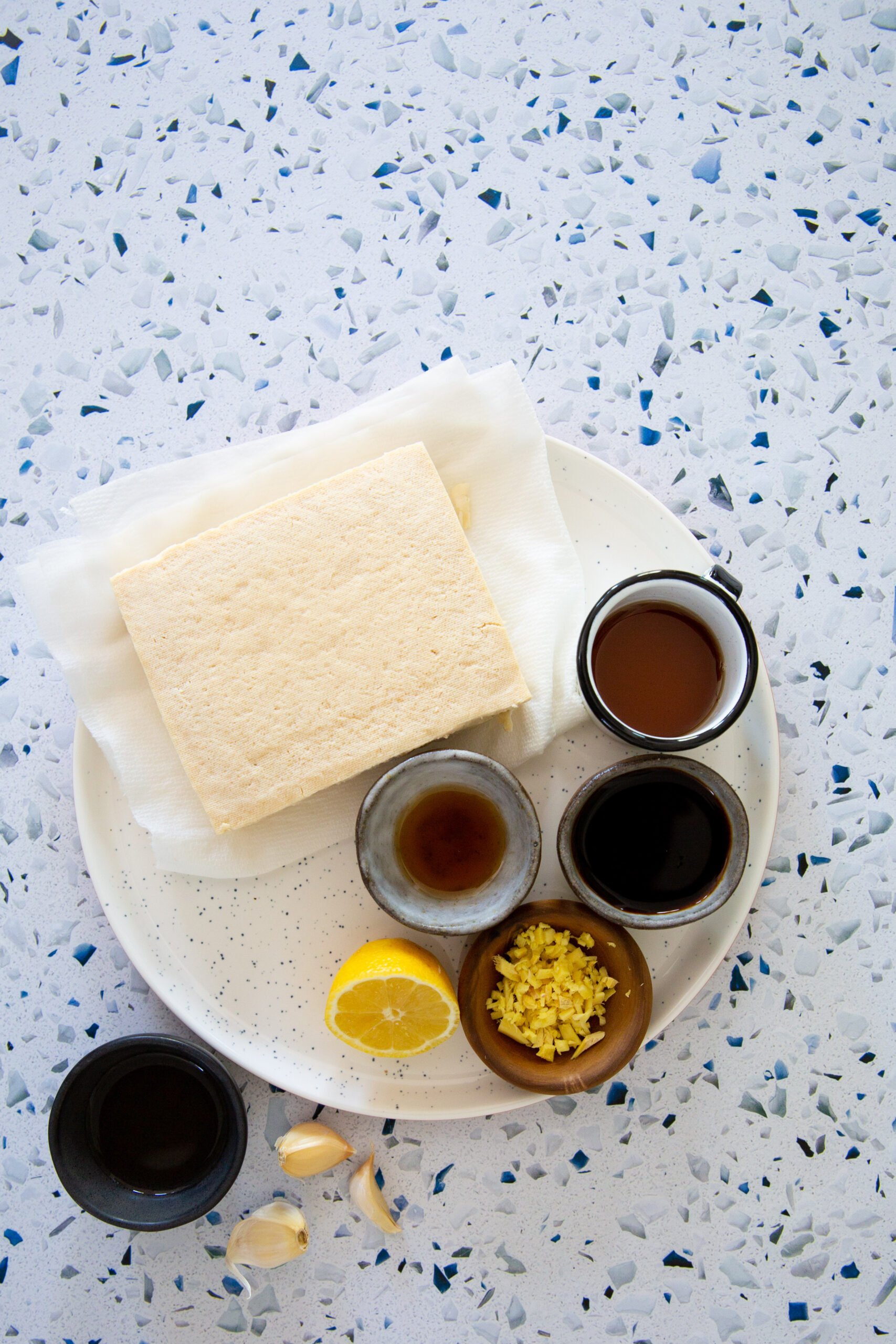 Un plato blanco sobre una encimera moteada contiene un bloque de tofu sobre toallas de papel, lo que recuerda a la preparación de ingredientes para un bánh mì. Alrededor del tofu hay pequeños cuencos que contienen salsa de soja, vinagre, jengibre rallado, dientes de ajo y medio limón, todo ello dispuesto en un semicírculo alrededor del tofu.