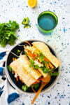 A bánh mì sandwich filled with pickled carrots, daikon, cilantro, and green chili is served on a black plate. A wooden spoon lies beside it. A green glass of water, sprigs of cilantro, a sliced green chili, and half a lemon are placed on the terrazzo countertop.