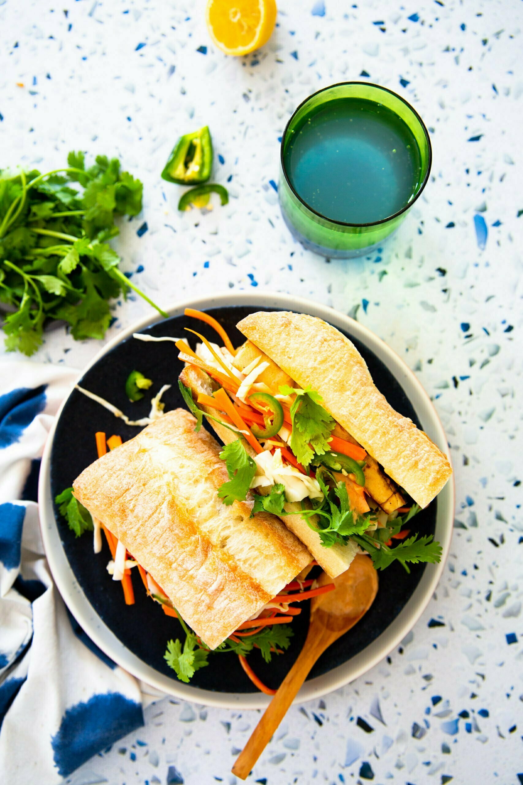 Un vibrante sándwich bánh mì en un plato negro hace alarde de verduras como zanahorias y cilantro envueltos en pan crujiente. Cerca, una cuchara de madera, un limón cortado por la mitad, hojas de cilantro, chile verde y un vaso verde lleno de líquido descansan sobre una superficie de terrazo, elegantemente envuelta en un paño azul y blanco.