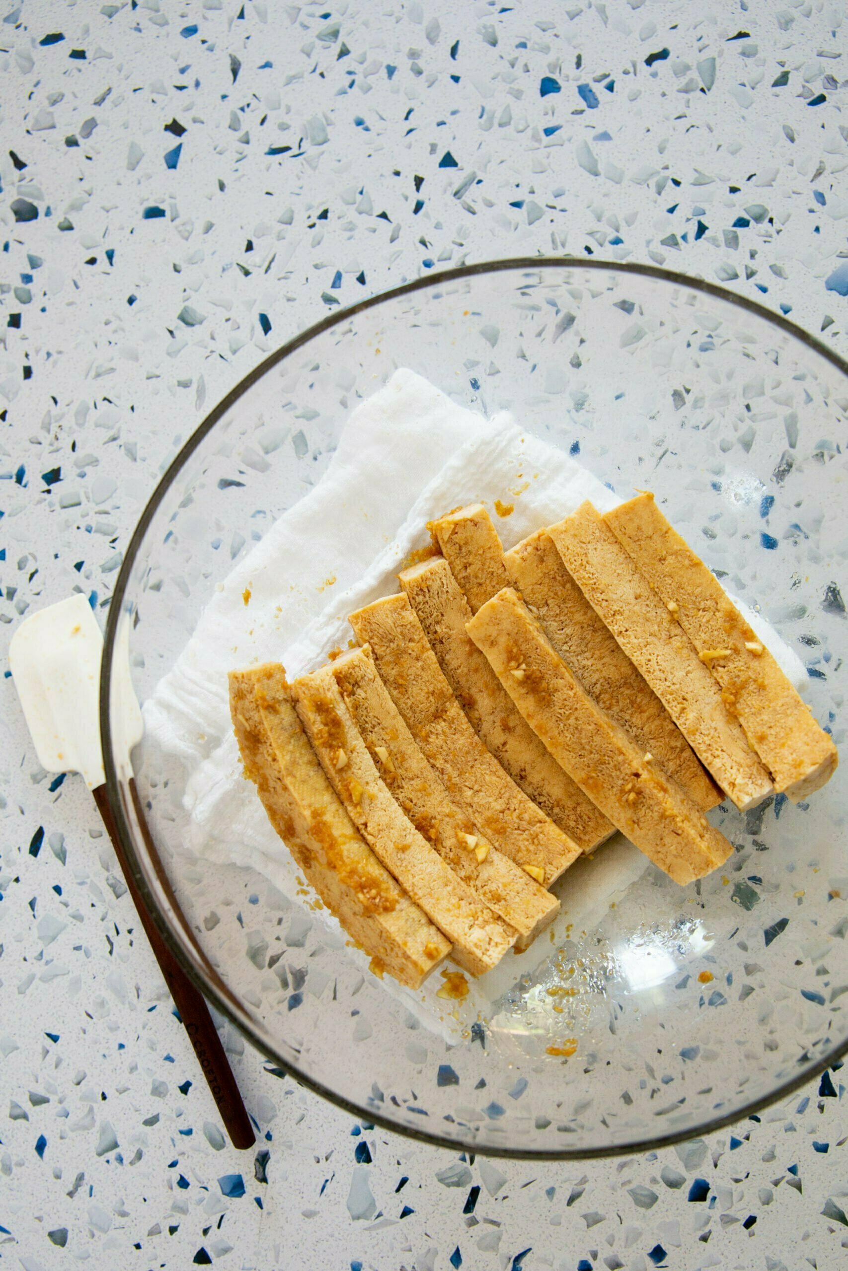 Un bol de vidrio transparente sobre una encimera moteada de blanco y gris contiene trozos de masa de galleta en rodajas, que recuerdan la delicada textura del bánh mì, apilados cuidadosamente sobre un paño blanco. Una espátula blanca con un mango oscuro reposa al lado del bol. La masa de color marrón claro se desmorona y está lista para hornear.