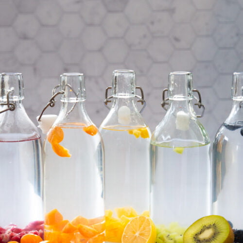 Five glass bottles filled with infused water are lined up in front of a hexagonal tile background. Each bottle features fruits like raspberries, kumquats, and mango, reminiscent of the best flavored lemonade. Assorted fresh fruits are scattered in front of the bottles, adding a vibrant touch.