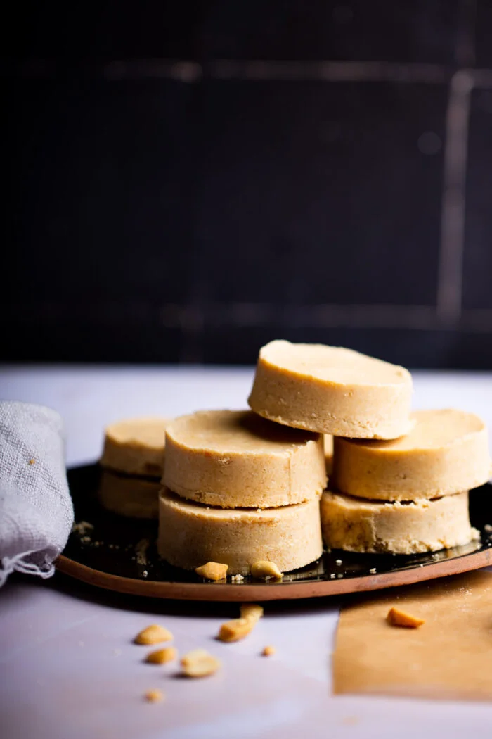 A stack of five round, beige cookies is on a dark plate against a black background. They resemble crumbly and rich mazapán, with cookie crumbs scattered on the white surface. To the side, a light gray cloth adds texture to the scene.