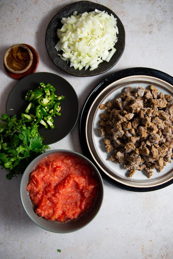 cebolla picada, cilantro, jalapeño en rodajas y carne vegana fozen en platos junto a un bol de puré de tomate
