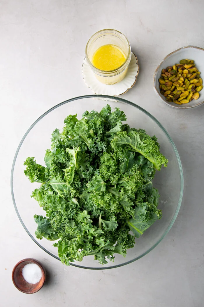 hojas de kale en una ensaladaeera, aderezo en un frasco y pistaches en un tazón.