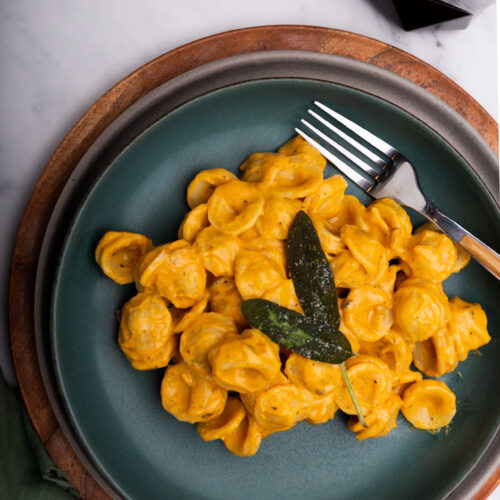 Pasta vegana cremosa de calabaza coronada con una ramita de salvia, servida con tenedor.