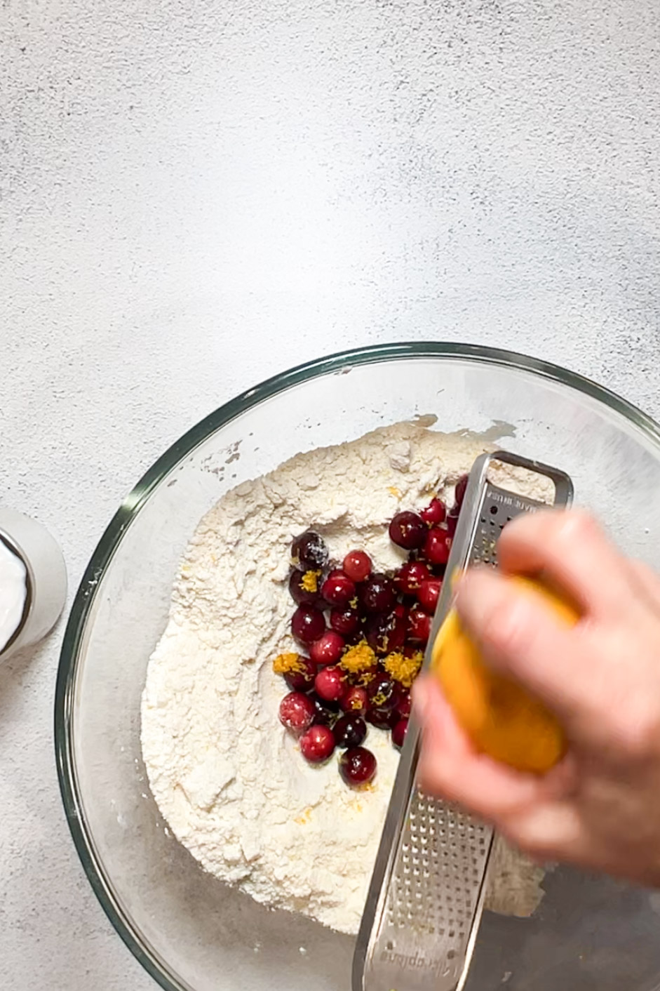 A person preparing vegan cranberry orange scones by sifting flour and cranberries into a bowl.
