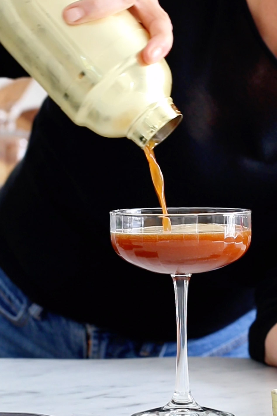 A woman preparing a shaken carajillo by pouring a drink into a glass.