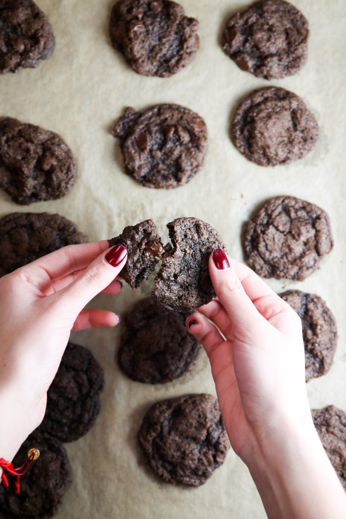 Unas manos con esmalte de uñas rojo parten una galleta de chocolate por la mitad sobre una bandeja para hornear cubierta con papel de pergamino. La bandeja contiene varias galletas de doble chocolate, dispuestas en filas. Las galletas tienen una apariencia texturizada y desmenuzable.