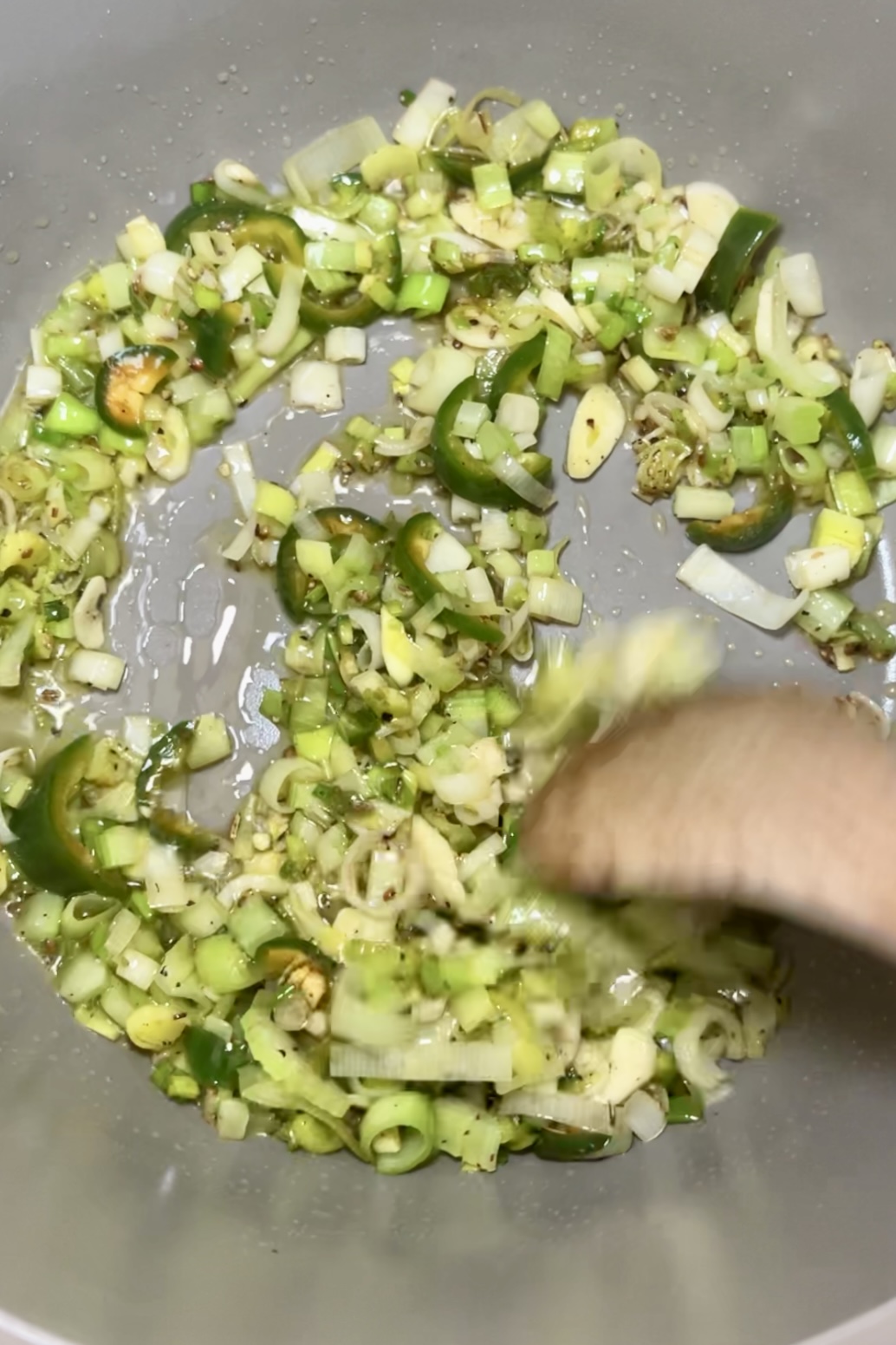 Una imagen de primer plano de puerros verdes picados y ajo salteados en una sartén plateada, con burbujas de aceite visibles, para una receta de Minestrone Verde. Una cuchara de madera está removiendo la mezcla.