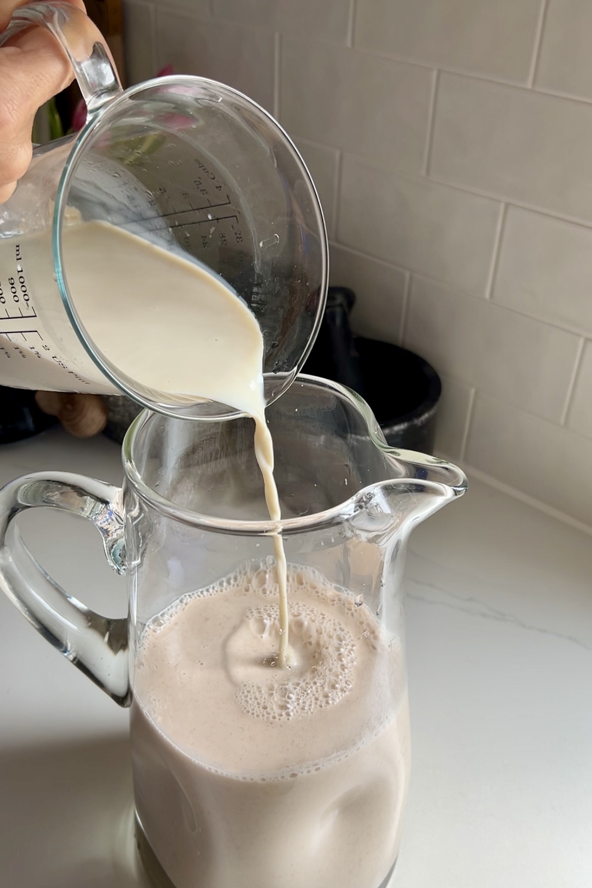 Una persona vierte un chorro de leche cremosa de una taza medidora en una jarra de cristal llena de espumosa horchata vegana de color marrón claro, colocada contra el salpicadero de una cocina.