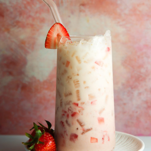 Una refrescante horchata de fresa en un vaso transparente, lleno de hielo y fresas troceadas, coronado con una fresa entera en el borde. La bebida se sirve con una pajita de cristal, en un plato ligero