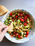 Se muestra una mano mojando un trozo de tortilla de maíz en una colorida ensalada vegana de ceviche en un cuenco.
La ensalada consiste en garbanzos, tomates cherry, rodajas de pepino, aceitunas y verduras, todo mezclado con un aliño ligero.
El cuenco está moteado de azul y blanco.
Cerca hay otro trozo de tortilla.