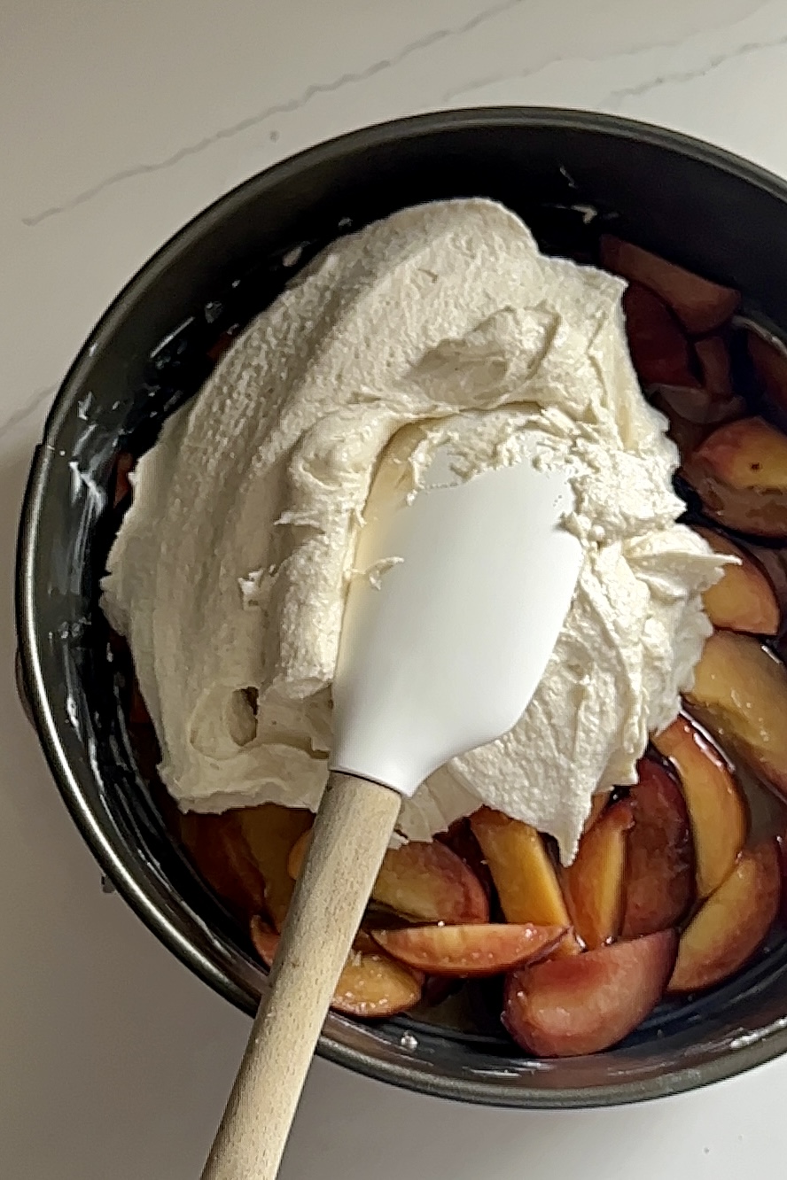 Un molde desmontable lleno de duraznos en rodajas (durazno) se cubre con una cucharada de masa blanca cremosa. Una espátula blanca con un mango de madera clara esparce la mezcla uniformemente sobre las frutas, preparando este delicioso volteado pastel para hornear. El fondo es una encimera blanca inmaculada.