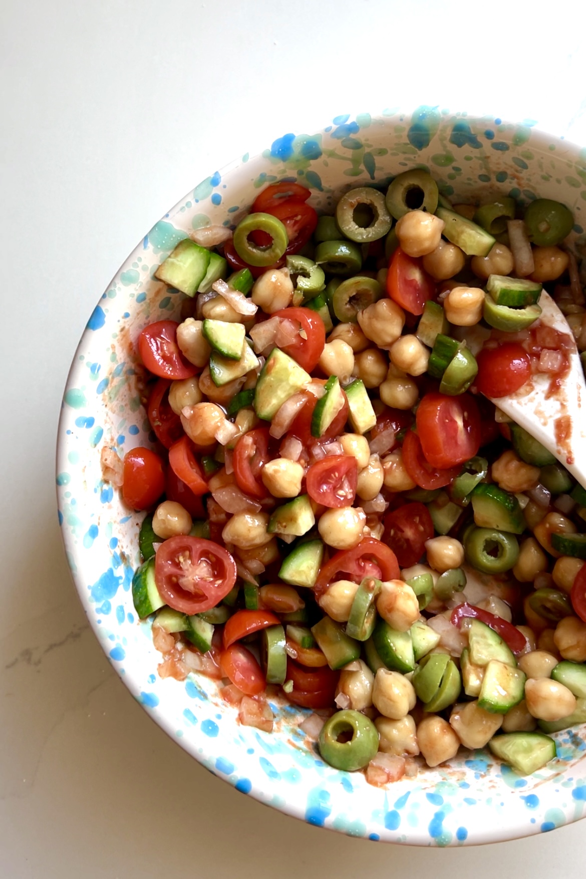 Una vibrante ensalada vegana en un cuenco moteado de blanco.
La ensalada de garbanzos se compone de tomates cherry cortados por la mitad, pepinos picados, aceitunas verdes cortadas en rodajas y cebollas rojas cortadas en rodajas finas.
Ligeramente aliñada y mezclada, con una cuchara mezcladora blanca dentro del cuenco.
El fondo es una superficie clara.   