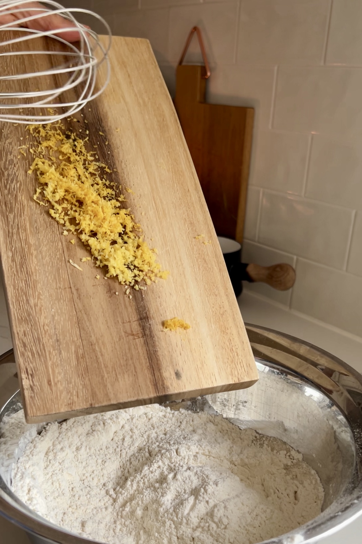 Primer plano de cáscara amarilla rallada sobre una tabla de cortar de madera que se añade a un cuenco metálico que contiene harina. Al fondo se ve una batidora metálica, y también parte de un alicatado de cocina. La escena capta un paso de la preparación de ingredientes en el proceso de horneado de Muffins veganos de limón y arándanos hechos desde cero.