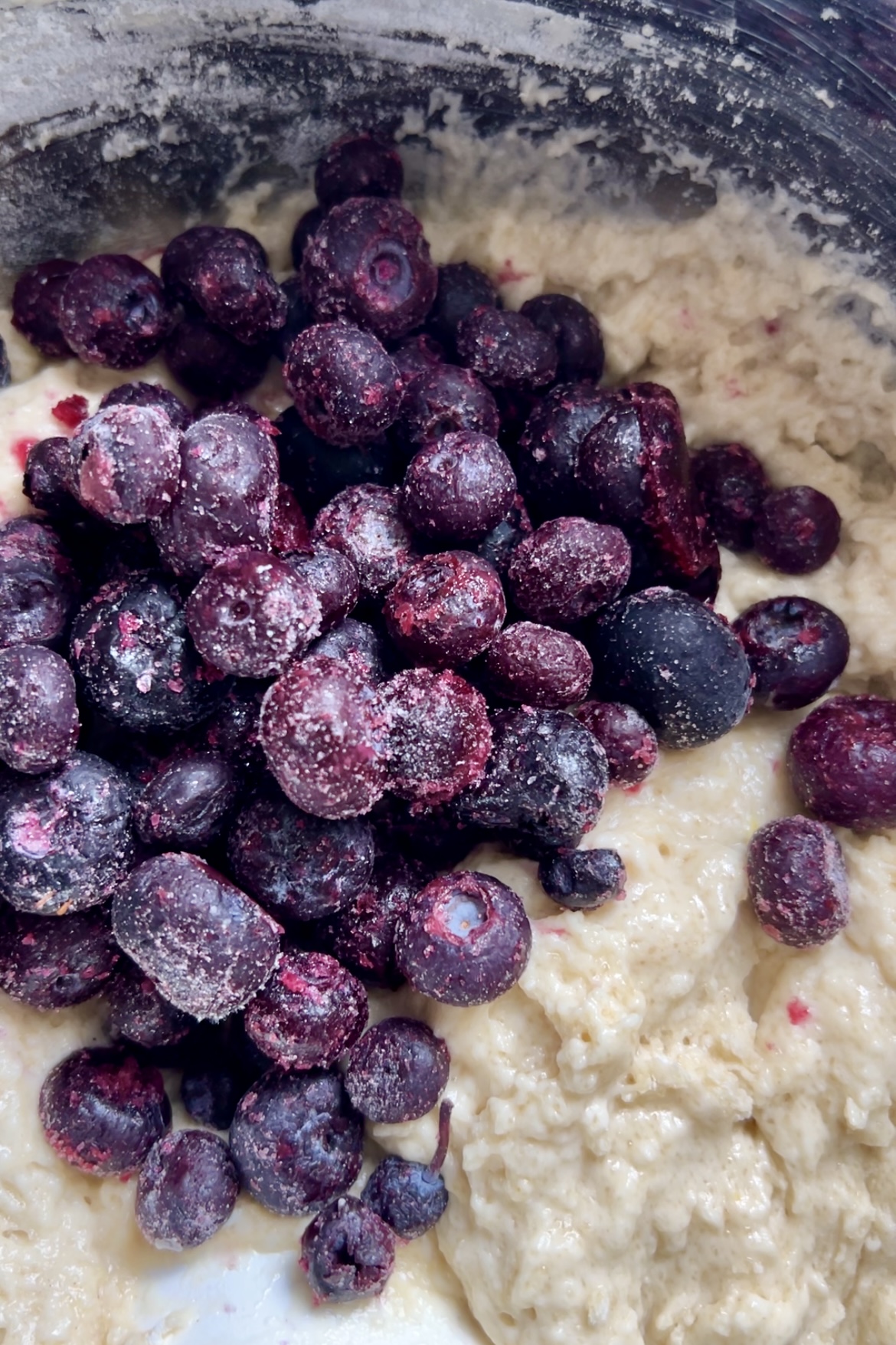 Un primer plano de una mezcla cremosa y espesa con avena y una generosa cantidad de arándanos congelados. Los arándanos, cubiertos de escarcha para darles un aspecto ligeramente blanco y moteado, aportan un profundo contraste púrpura a la avena de color claro, perfecto para esos Muffins de limón y arándanos hechos desde cero o incluso como delicia vegana.