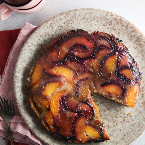 Un pastel volteado glaseado reposa sobre un plato moteado de color beige, al que le falta una rebanada. El pastel presenta rebanadas de durazno de color naranja vibrante y ciruela morada sobre una superficie caramelizada. A su lado hay dos tenedores vacíos, una servilleta roja y tazas de color rojo oscuro apiladas, que proyectan sombras sutiles sobre la encimera clara.