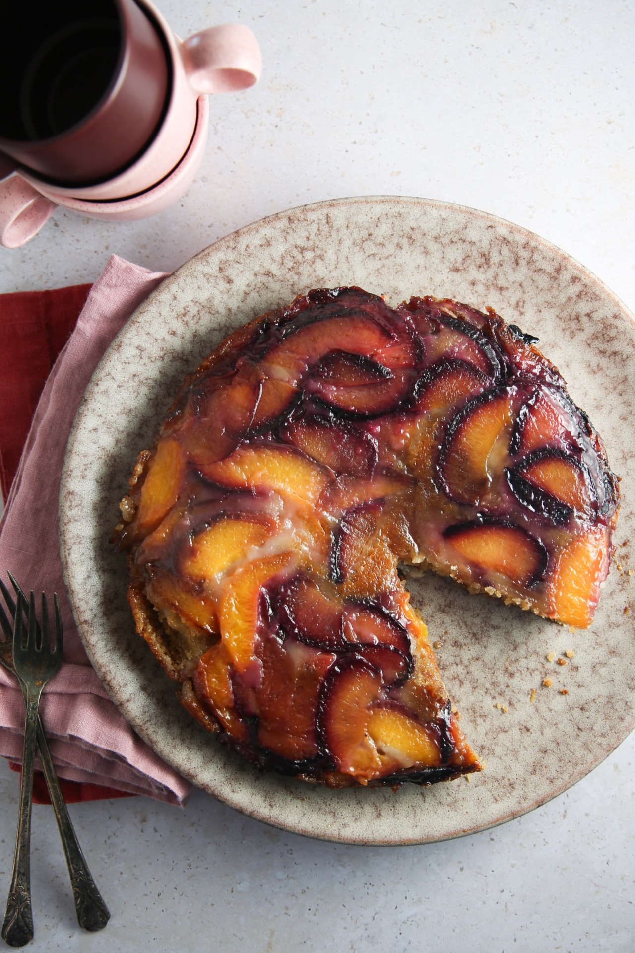 Un pastel volteado glaseado reposa sobre un plato moteado de color beige, al que le falta una rebanada. El pastel presenta rebanadas de durazno de color naranja vibrante y ciruela morada sobre una superficie caramelizada. A su lado hay dos tenedores vacíos, una servilleta roja y tazas de color rojo oscuro apiladas, que proyectan sombras sutiles sobre la encimera clara.