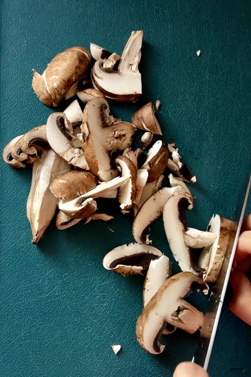 A person's hand is seen slicing brown mushrooms on a dark green cutting board using a knife. The mushrooms are in various stages of being cut, with some already sliced and others whole or halved. The focus is on the cutting process and the texture, perfect for preparing a flavorful Mexican Mushroom Soup.