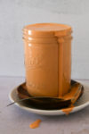 A jar filled with creamy, orange-colored chipotle crema overflows slightly, with the sauce dripping down the side. The jar is placed on two stacked saucers, one dark and one light. A spoon with sauce remnants lies across the saucers. The white background enhances the sauce's vibrant color.