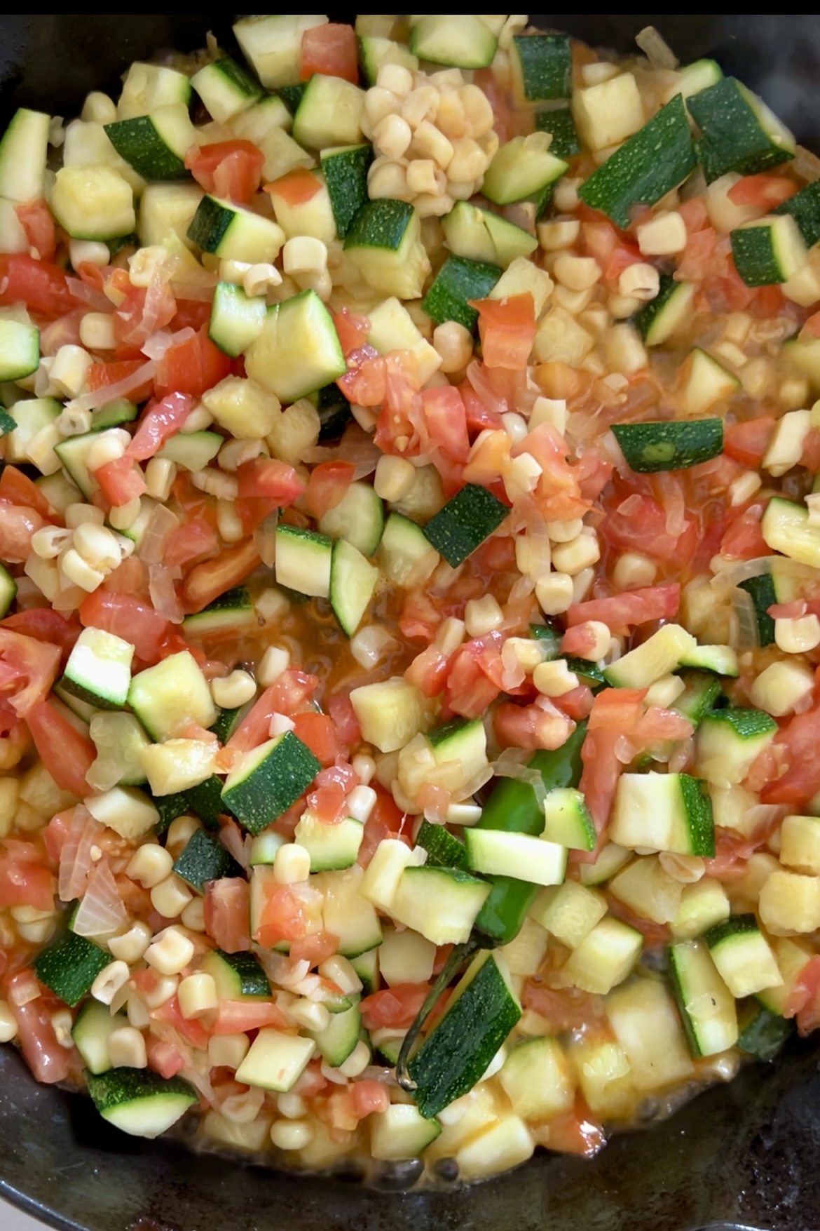 Un primer plano muestra una colorida mezcla de dados de calabacín, maíz, tomates y cebollas salteados en una sartén.
Esta receta tradicional mexicana, conocida como Calabacitas, muestra verduras frescas y vibrantes, con el calabacín verde, el maíz amarillo, los tomates rojos y las cebollas translúcidas que crean un contraste visualmente atractivo. 