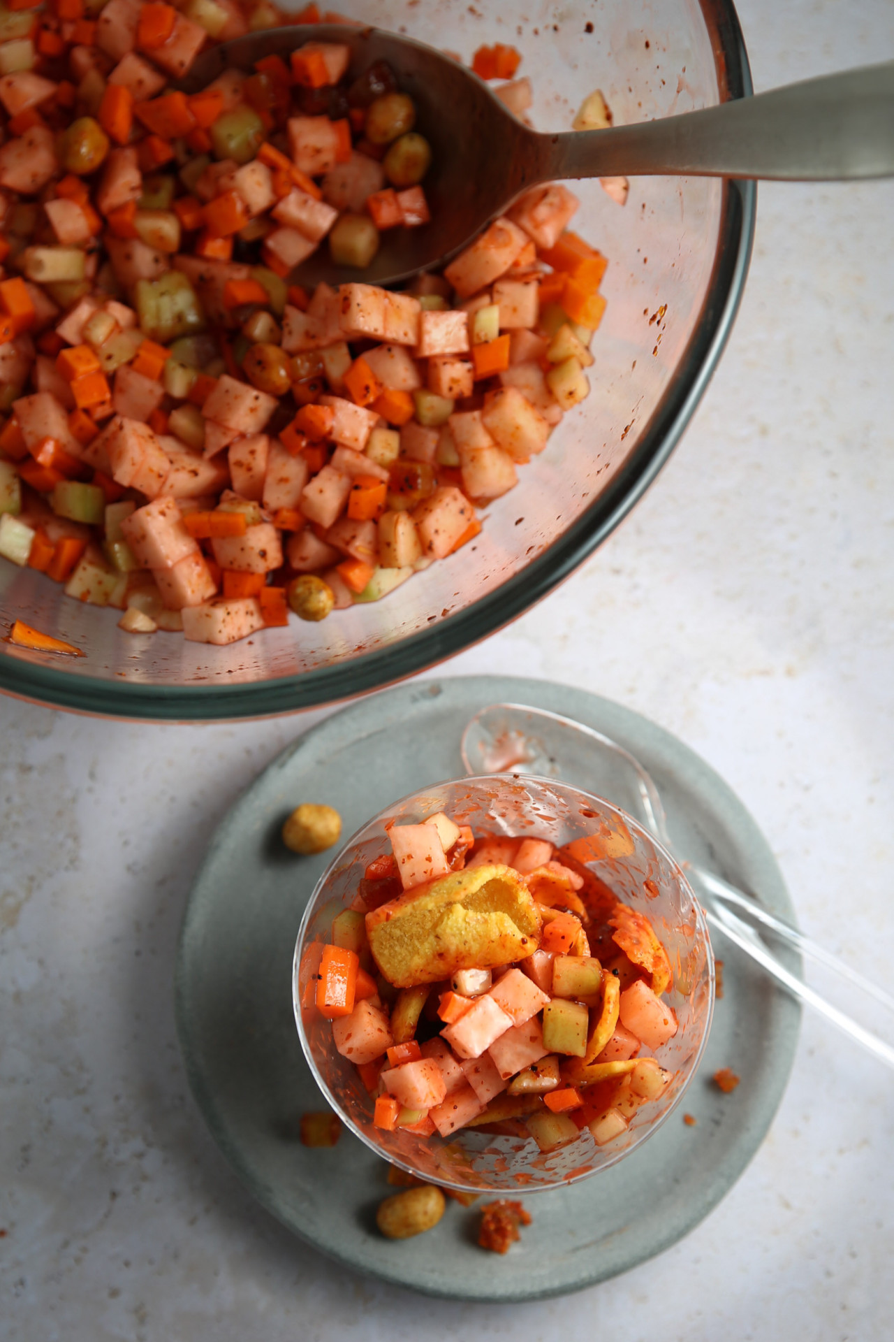 Primer plano de una ensalada mixta de verduras en un cuenco de cristal transparente sobre un plato gris claro con un tenedor transparente.
La ensalada incluye verduras cortadas en dados, como zanahorias, manzanas y pepinos, sazonadas con especias: un aperitivo de fiesta por excelencia.
Al fondo se ve un cuenco grande con la misma ensalada y una cuchara de servir.  