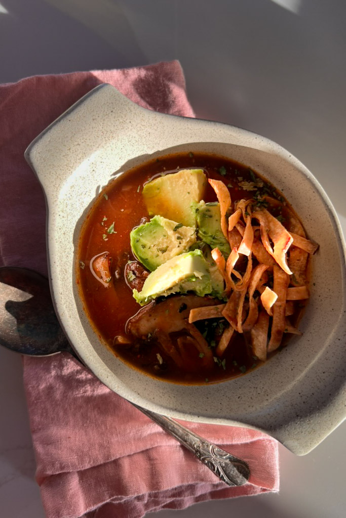 Un plato de sopa de tortilla relleno con un rico caldo rojo, cubierto con rodajas de aguacate, tiras de tortilla crujientes, pollo desmenuzado y hierbas. El cuenco reposa sobre una servilleta de tela rosa, con una cuchara metálica visible en un lateral. Esta abundante comida se parece al tradicional caldo de chipotle. El entorno está iluminado por luz natural.