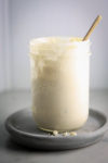 A creamy substance in a mason jar with a spoon sticking out is placed on a round gray ceramic plate. The plate has crumbs of the Vegan Blue Cheese Dressing around the base of the jar. The neutral gray background and excellent lighting provide a clear view of this delectable vegan dressing recipe.