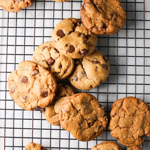 Una hornada de galletas de chocolate se enfría sobre una rejilla negra.
Las galletas doradas, con un toque de especias de calabaza, tienen trocitos de chocolate visibles y la superficie ligeramente agrietada.
Una galleta está partida por la mitad, y la disposición general es informal, con algunas galletas superpuestas de forma irregular sobre la rejilla.