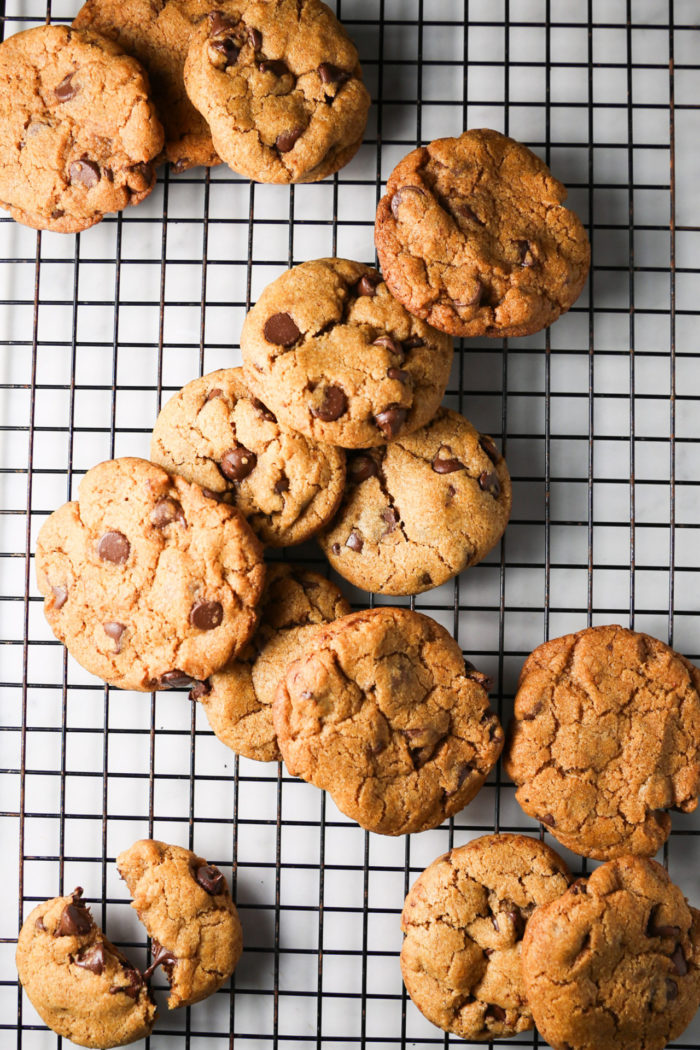 Una hornada de galletas de chocolate se enfría sobre una rejilla negra.
Las galletas doradas, con un toque de especias de calabaza, tienen trocitos de chocolate visibles y la superficie ligeramente agrietada.
Una galleta está partida por la mitad, y la disposición general es informal, con algunas galletas superpuestas de forma irregular sobre la rejilla.