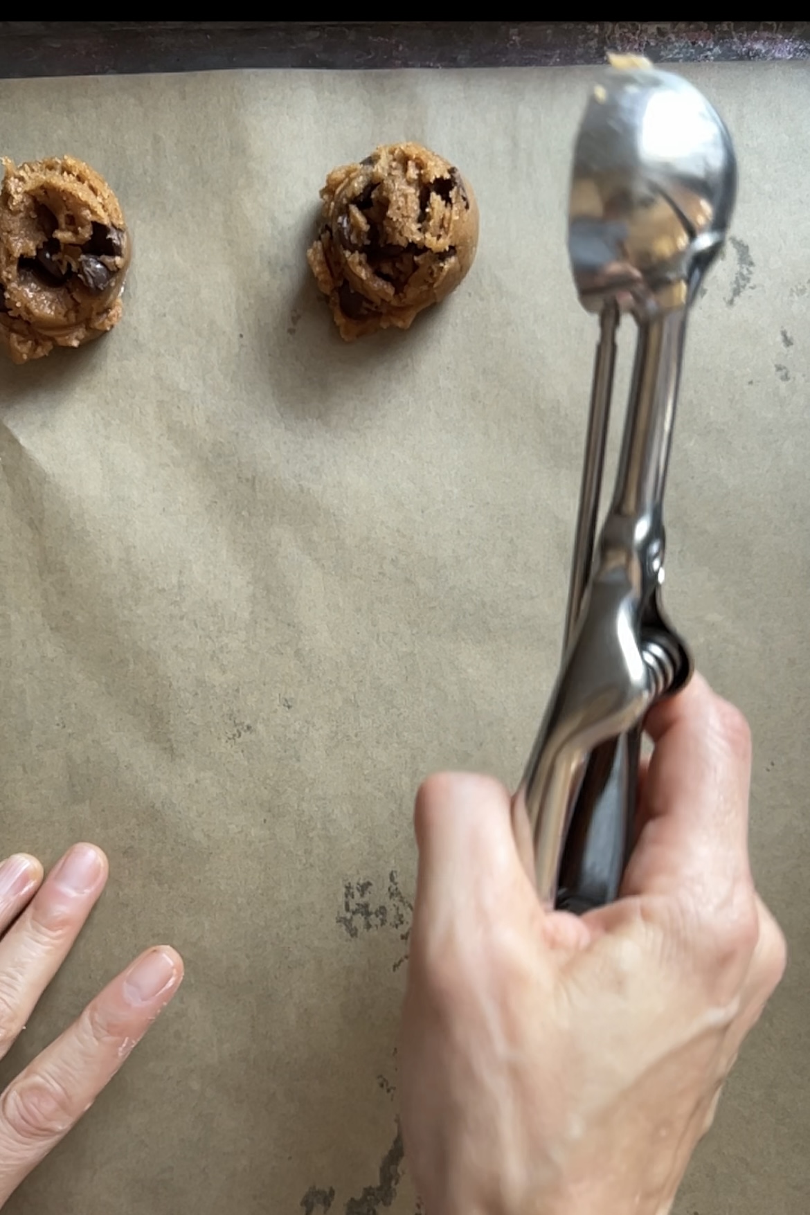 Una mano vierte la masa de galletas con pepitas de chocolate con una cuchara metálica para galletas, colocando las bolas de masa en una bandeja para hornear forrada con papel pergamino.
Dos bolas de masa con especias de calabaza ya están en la bandeja, listas para hornear.
El fondo incluye una parte de una bandeja de horno de borde oscuro.  